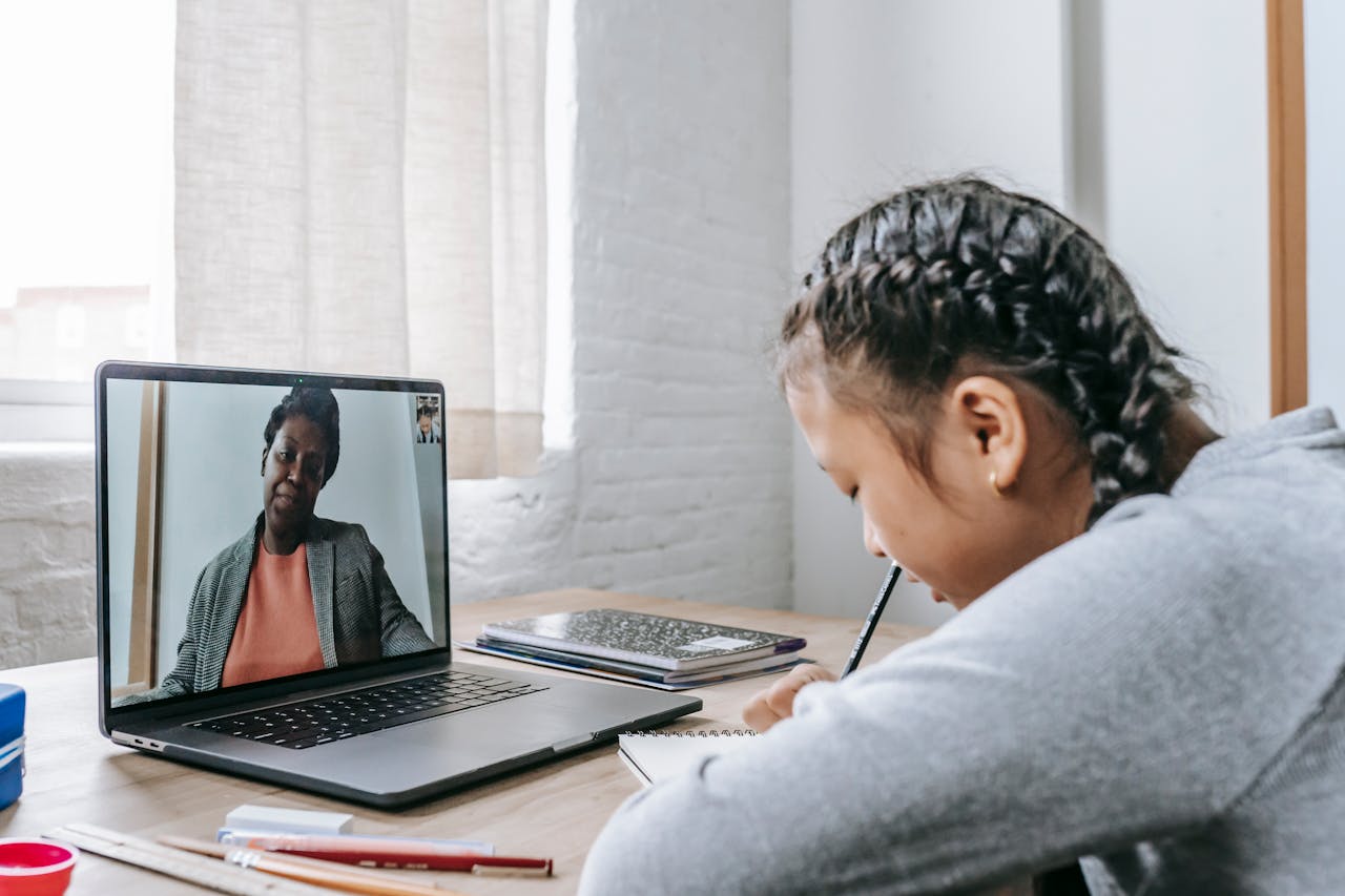 Side view of focused Asian schoolgirl sitting at wooden table and writing notes during remote lesson on laptop with African American teacher