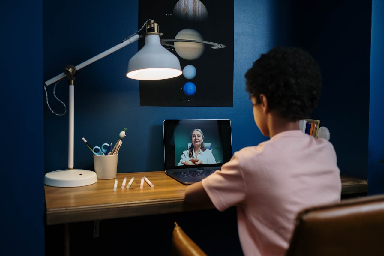 A young student engaged in an online class with a teacher, sitting at a desk with a laptop.